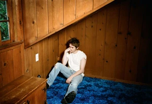 Actor Austin Abrams seated on a blue carpet in a wood-paneled room wearing a white t-shirt and light blue jeans
