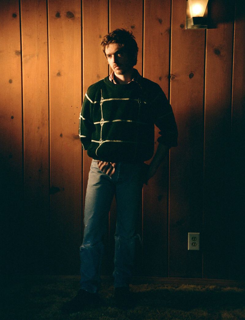 Austin Abrams posing against a vertical wood panel wall wearing a dark green patterned sweater and denim jeans.