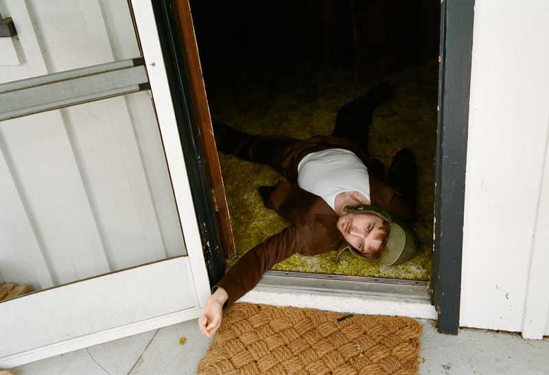 Austin Abrams lying on green carpet in a doorway wearing a brown jacket and white t-shirt for HERO Magazine.