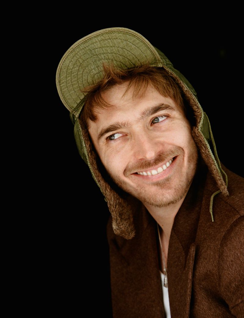 Portrait of actor Austin Abrams wearing an olive green trapper hat and brown jacket against a black background.