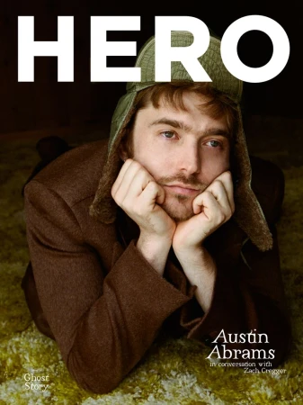 Austin Abrams resting his chin on his hands, wearing a brown jacket and green trapper hat on a textured rug