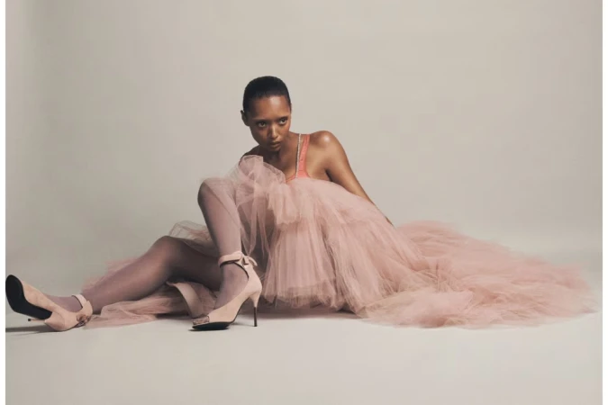Model Sophie Wilde wearing a voluminous pink tulle dress and high heels seated on a seamless backdrop