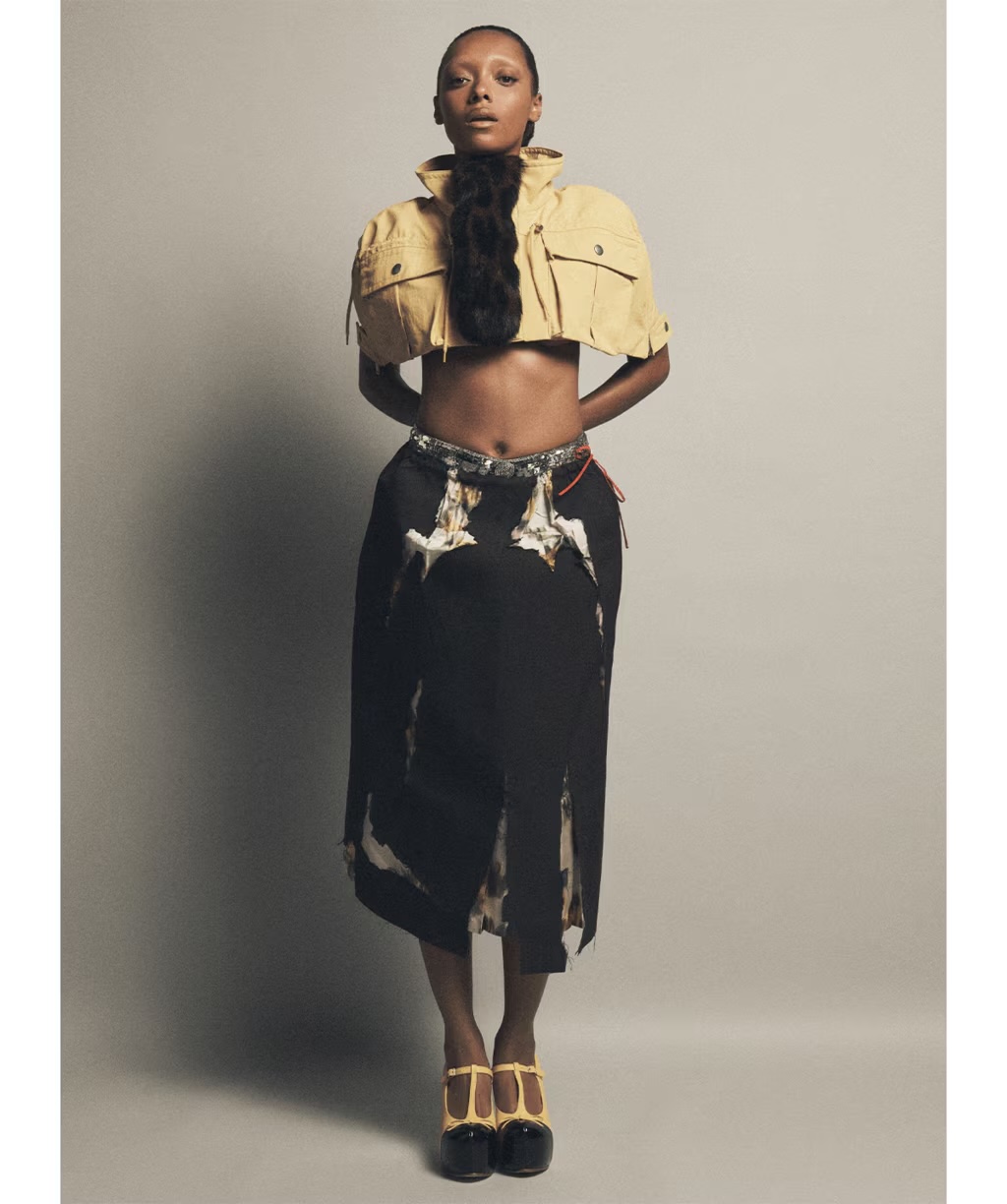 Sophie Wilde in a cropped yellow jacket and dark patterned skirt against a grey seamless studio background.