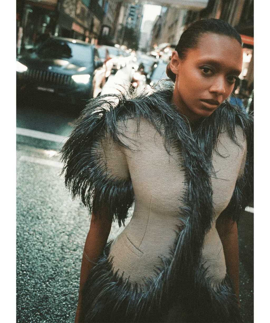Sophie Wilde in a structured beige garment with dark feathered trim standing on an urban street.