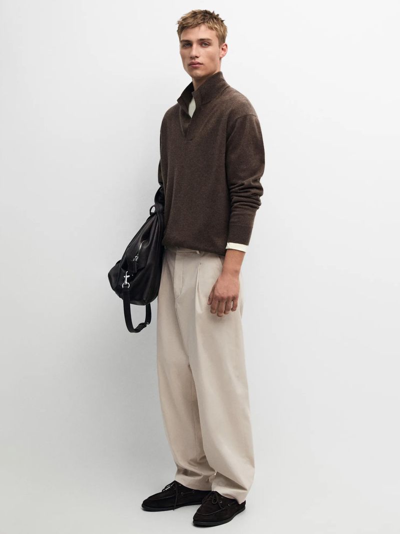 Young man standing against a white backdrop wearing a brown zip-collar sweater, light trousers, holding a black bag