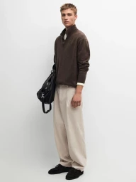 Young man standing against a white backdrop wearing a brown zip-collar sweater, light trousers, holding a black bag