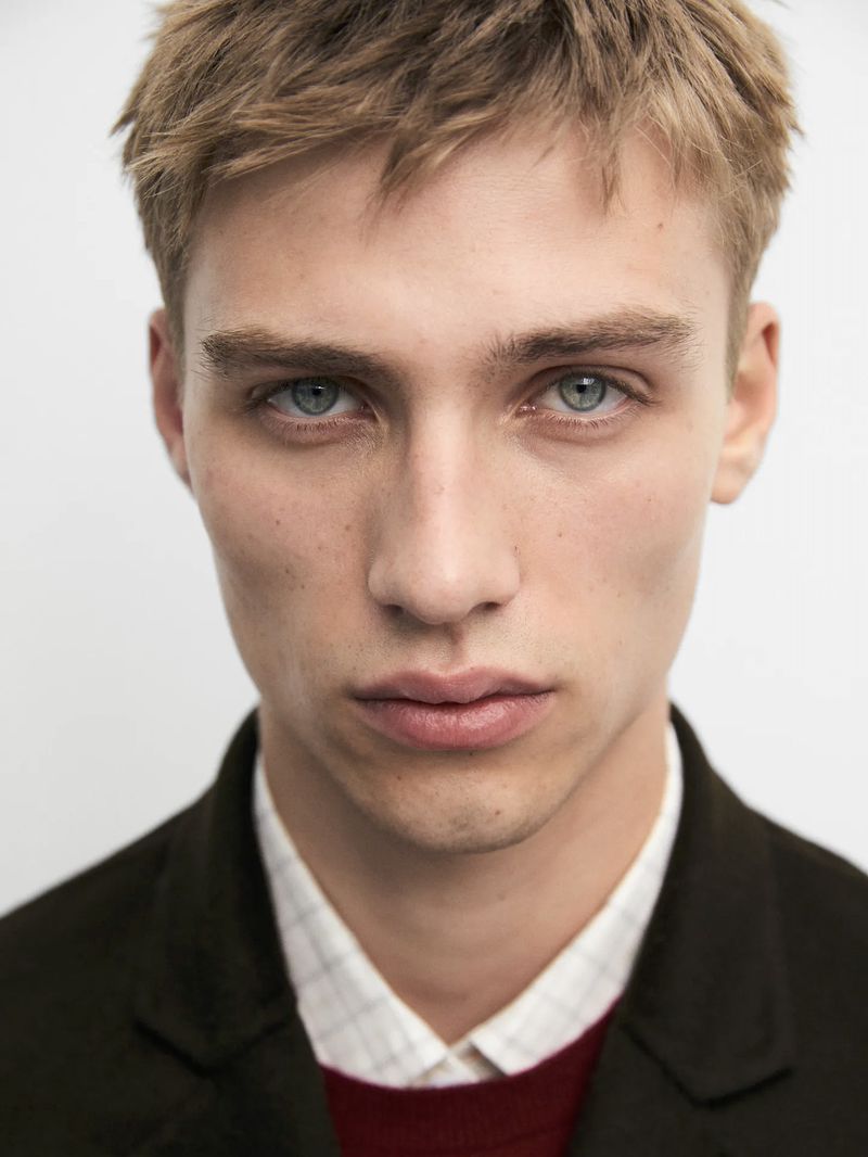 Close-up portrait of a young man with blonde hair and blue eyes wearing a dark jacket over a plaid shirt.