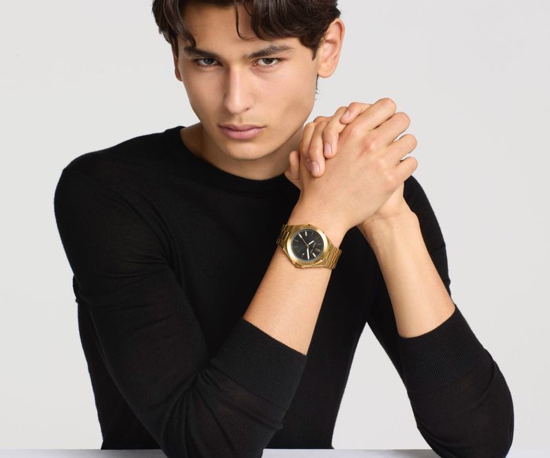 Model Ellis Mueller wearing a black crewneck sweater and gold wristwatch for Michael Kors against a plain white background