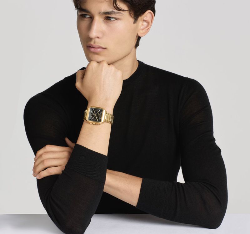 Close-up portrait of a male model wearing a black sweater and a gold chronograph watch against a plain grey backdrop.