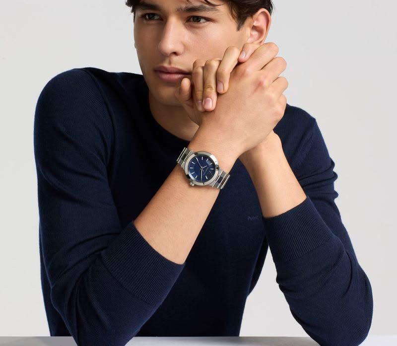 Model wearing a metal watch with a blue dial and a dark blue long-sleeve sweater against a white background