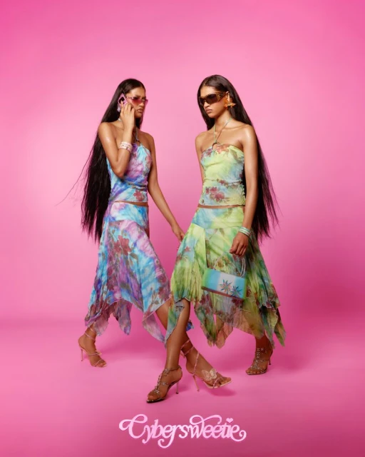 Two female fashion models in matching tie-dye outfits and sunglasses pose against a pink backdrop in a studio setting, holding a bag.