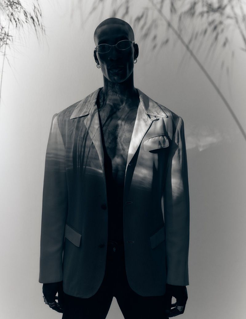 Black man wearing a blazer and sunglasses with linear shadow patterns cast across him against a light background.