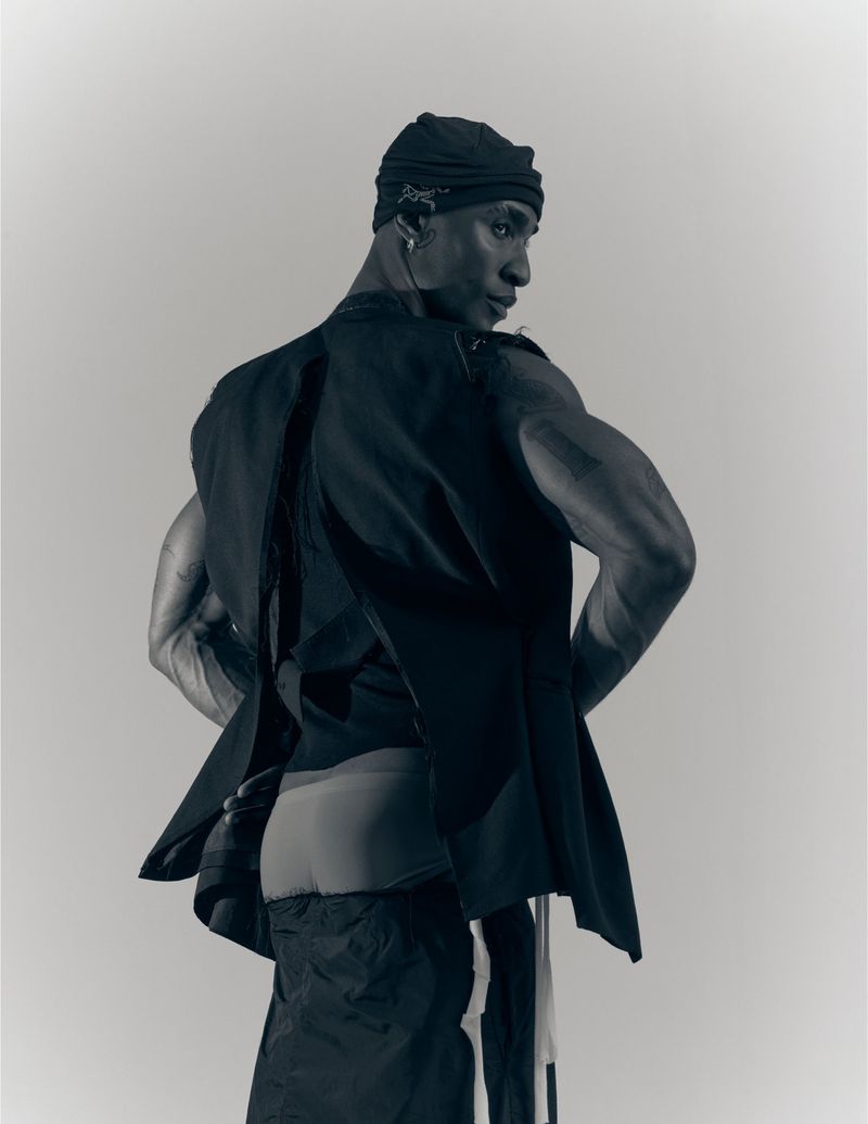 Man wearing a dark cap, black sleeveless vest and trousers looking over his shoulder against a plain grey background