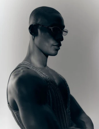 Portrait of a bald Black man wearing sunglasses and a textured sleeveless top against a plain light background.