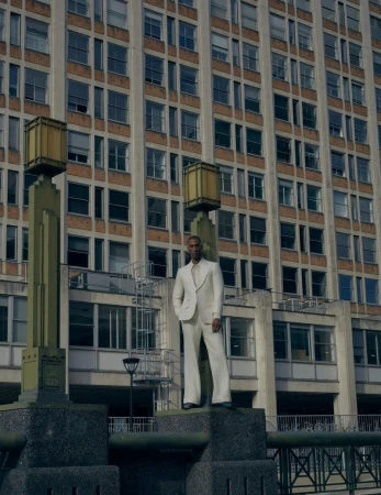 Man wearing a cream suit standing on a stone plinth in front of a large multi-story architectural building facade.