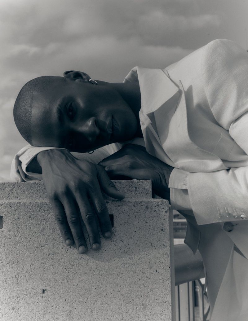 Portrait of a Black man in a white jacket leaning against a concrete block under a cloudy sky.