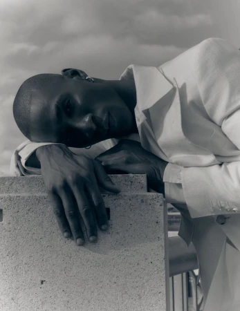 Portrait of a Black man in a white jacket leaning against a concrete block under a cloudy sky.