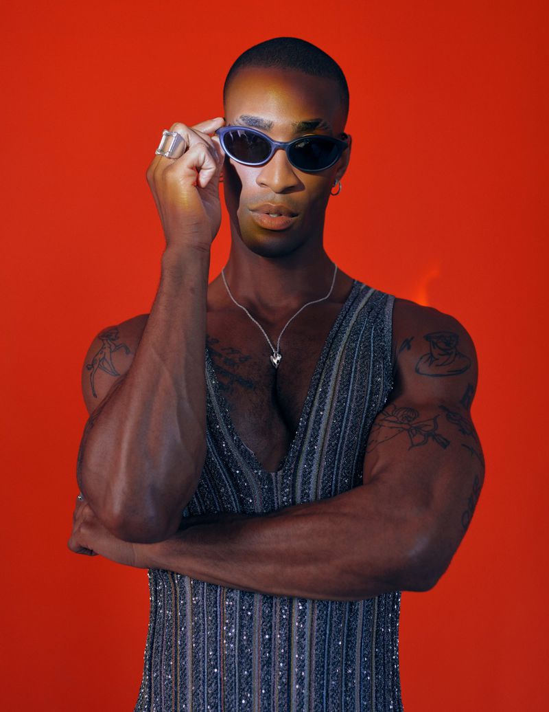 Black man wearing dark sunglasses and a glittery sleeveless top against a solid red background