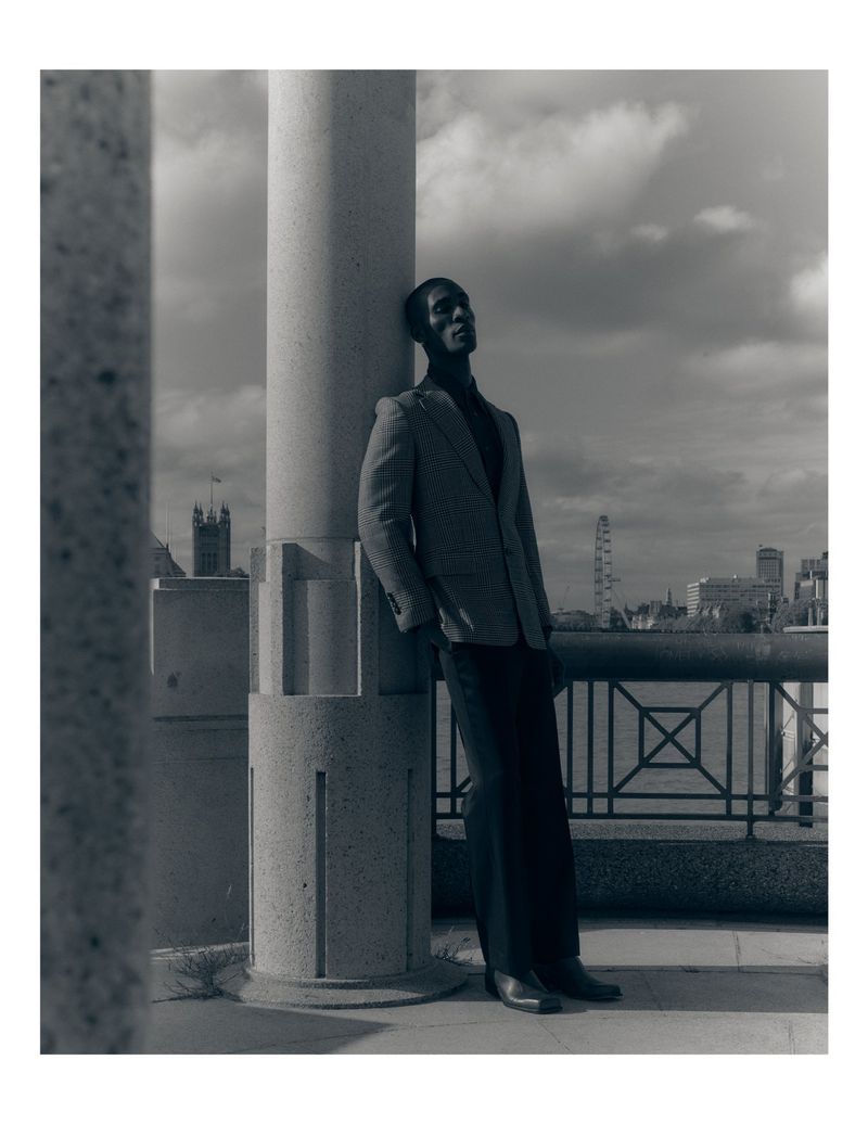 Black man wearing a blazer leaning against a large stone pillar outdoors against an urban skyline