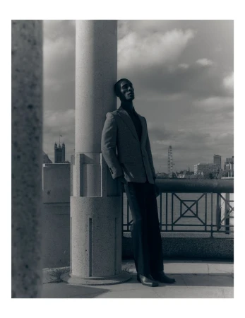Black man wearing a blazer leaning against a large stone pillar outdoors against an urban skyline
