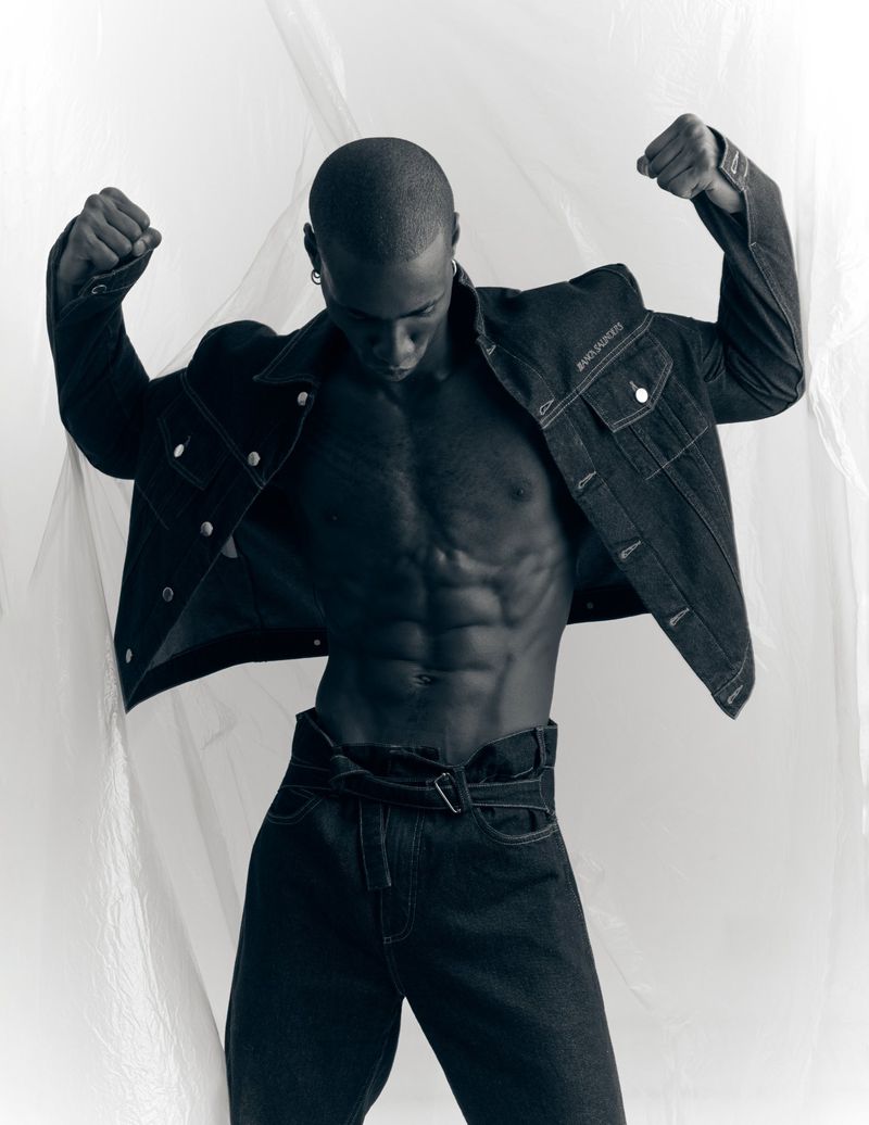 Black man with a muscular physique wearing a dark denim jacket and matching jeans posing against a translucent plastic backdrop