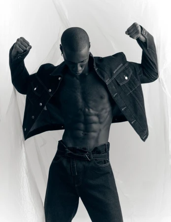 Black man with a muscular physique wearing a dark denim jacket and matching jeans posing against a translucent plastic backdrop