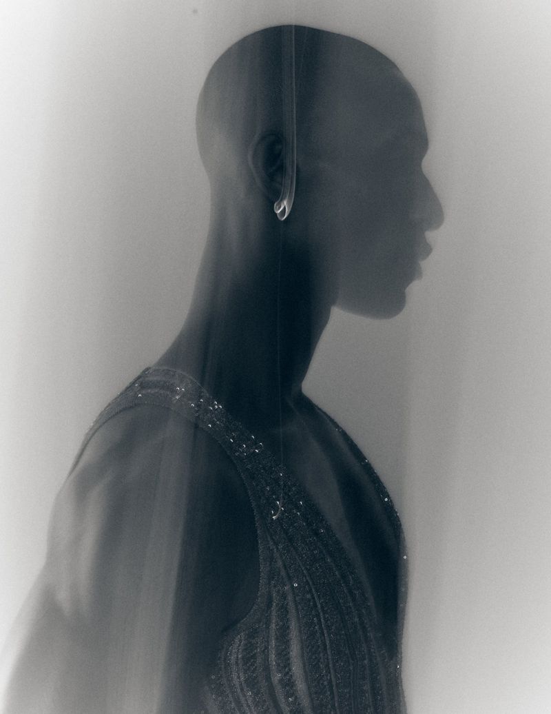 Profile portrait of a bald Black man wearing a beaded tank top, captured in a soft hazy monochrome style for Fucking Young.
