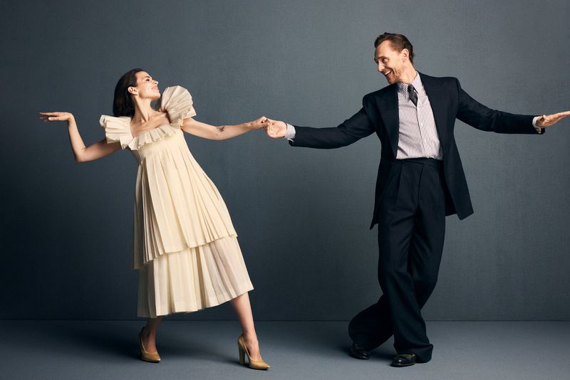 Tom Hiddleston in a suit and a woman in a cream ruffled dress holding hands against a grey seamless background