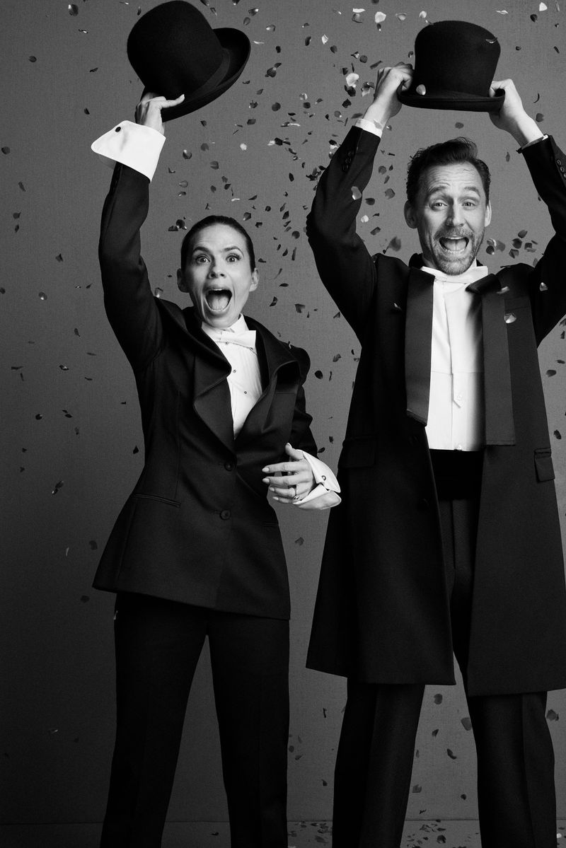 Actor Tom Hiddleston and co-star in black tie attire raising bowler hats amid falling confetti in a monochrome portrait.