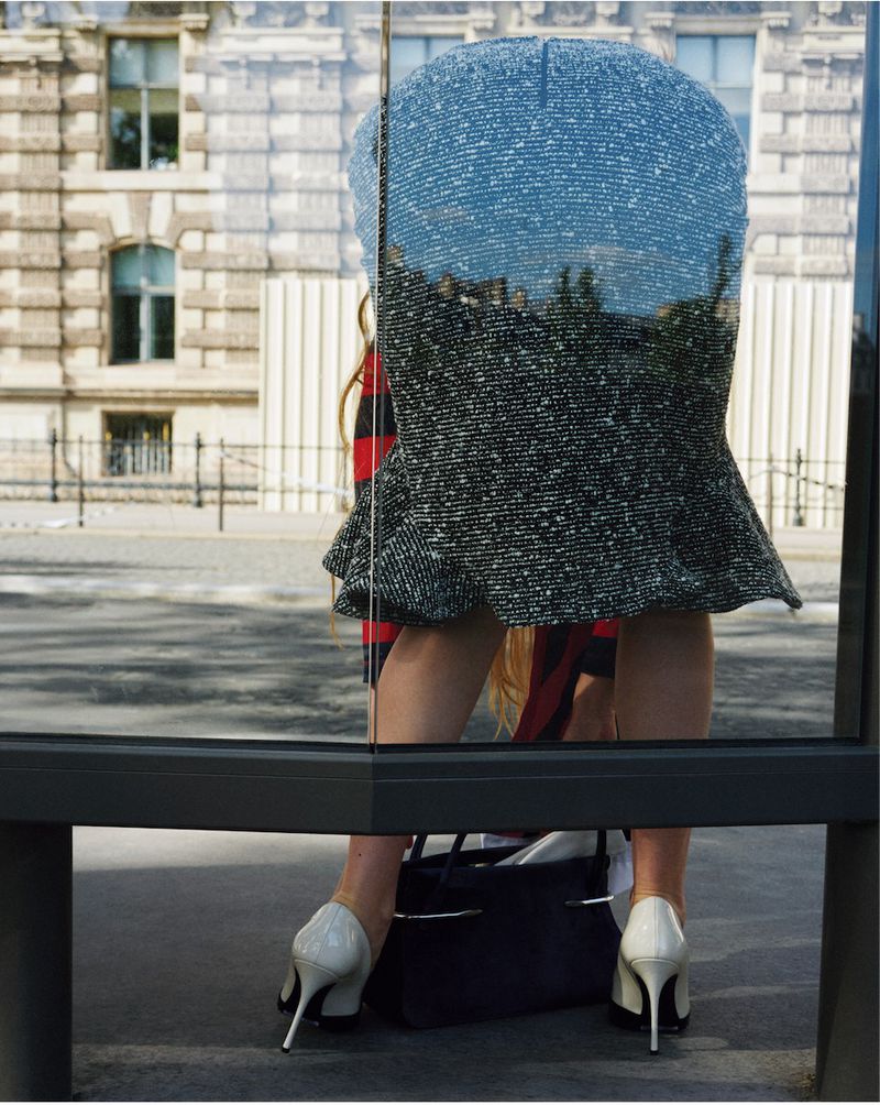A person viewed from behind wearing a textured tweed skirt, white stiletto heels, and holding a dark handbag near a glass bus stop.