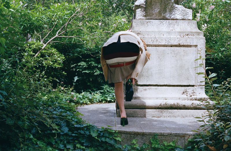 Woman bent over backwards against a stone monument in a lush garden setting wearing a blazer and skirt