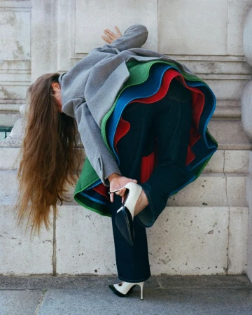 Model bending forward with long brown hair, wearing a grey jacket, flared skirt with green, blue, and red layers, jeans, and heels against stone walls.