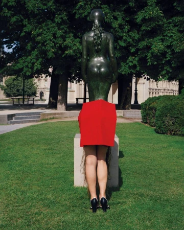 A person standing behind a bronze statue in a park, wearing a red garment over their lower body and heels