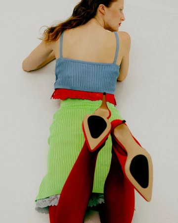 Model lying on a white studio background wearing a blue strap top, green knit skirt, and red heels.