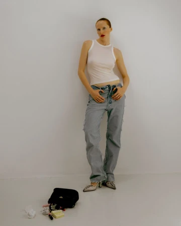 Model wearing a white tank top and light wash denim jeans standing against a plain white wall with a spilled handbag on the floor.