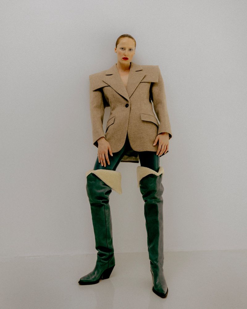 Model wearing structured tan blazer and green thigh high boots against a plain white studio background