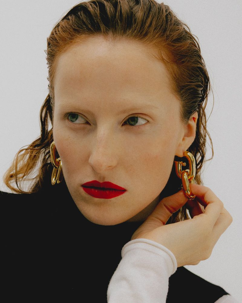 Close-up portrait of a woman with wet-look hair wearing bold red lipstick and chunky gold chain earrings against a white background.