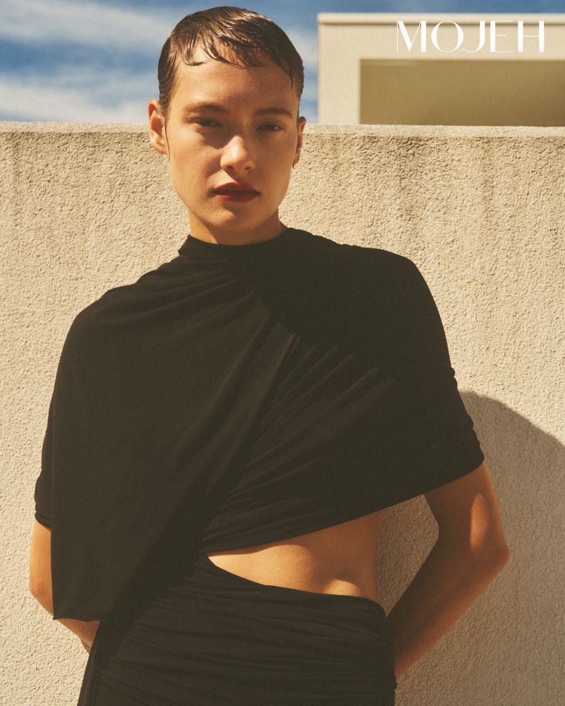 Model in black top standing against a textured wall under warm sunlight for Mojeh Magazine