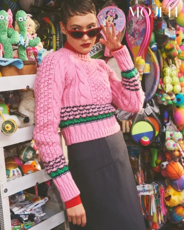 Model with short hair wearing red sunglasses and a pink cable knit sweater in front of a toy display