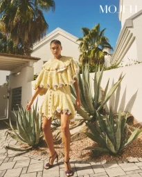 Model in ruffled yellow textured dress and heeled sandals standing outdoors among palm trees and agave plants.