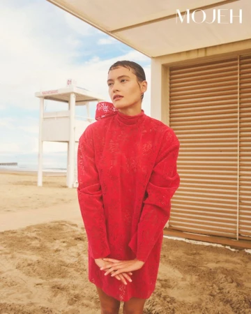 Model standing on a sandy beach wearing a vibrant red textured dress with a high collar and a large bow at the back.