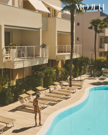Model standing by an outdoor swimming pool with lounge chairs and modern apartment buildings in the background