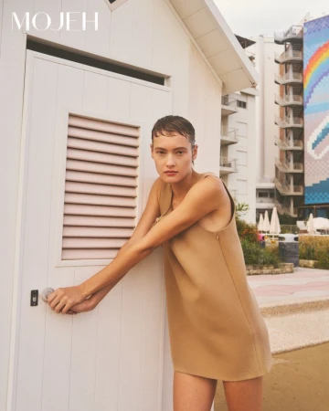 Model in camel sleeveless mini dress holding white door handle outdoors near building