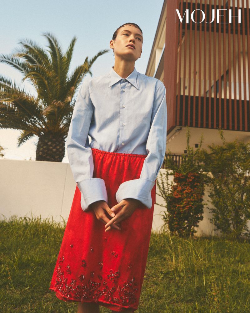Model standing outdoors wearing a light blue button-down shirt and a red skirt against a modern building and palm tree