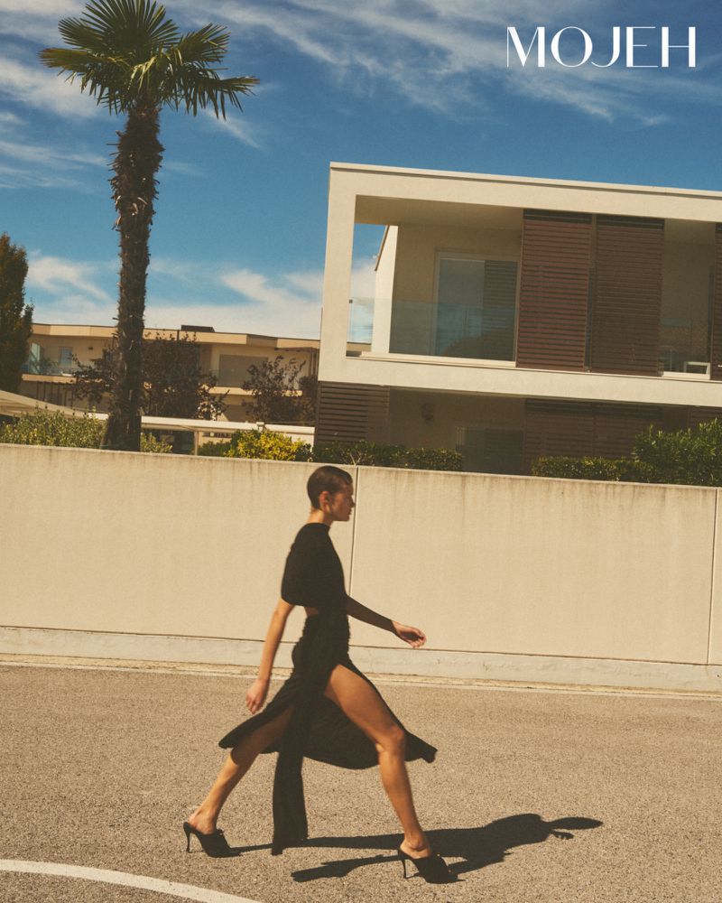 Model in a black dress walking outdoors near a modern building and palm tree for Mojeh Magazine