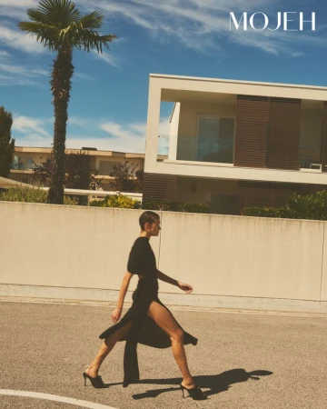 Model in a black dress walking outdoors near a modern building and palm tree for Mojeh Magazine