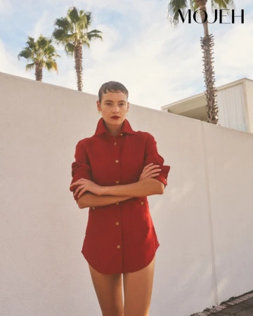 Model in red shirt with arms crossed posing in front of a white wall and palm trees