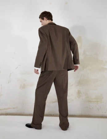 Model wearing a brown suit seen from behind against a textured white wall