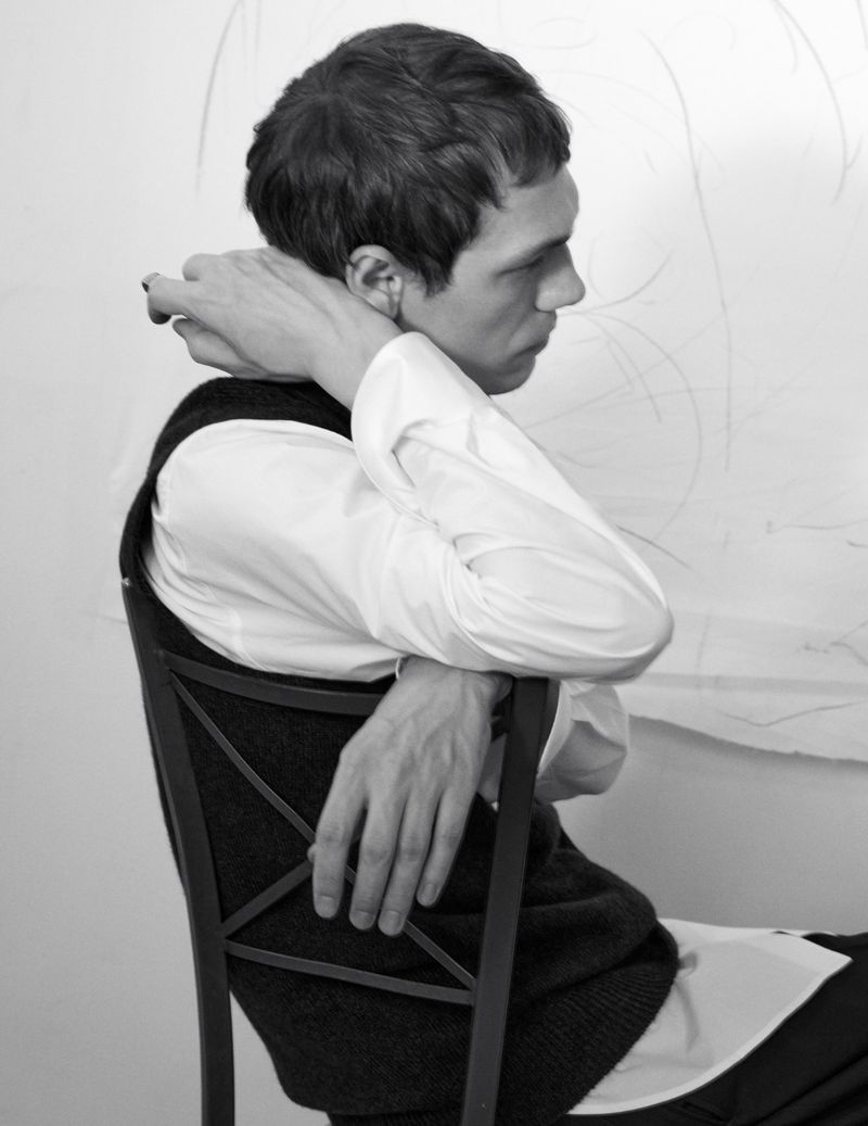 Model Aleksandr Gordeev seated backward on a chair in a white button-down shirt and dark vest