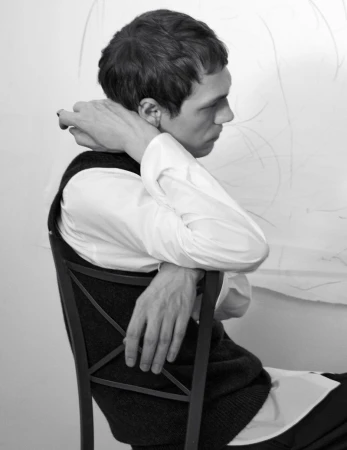 Model Aleksandr Gordeev seated backward on a chair in a white button-down shirt and dark vest